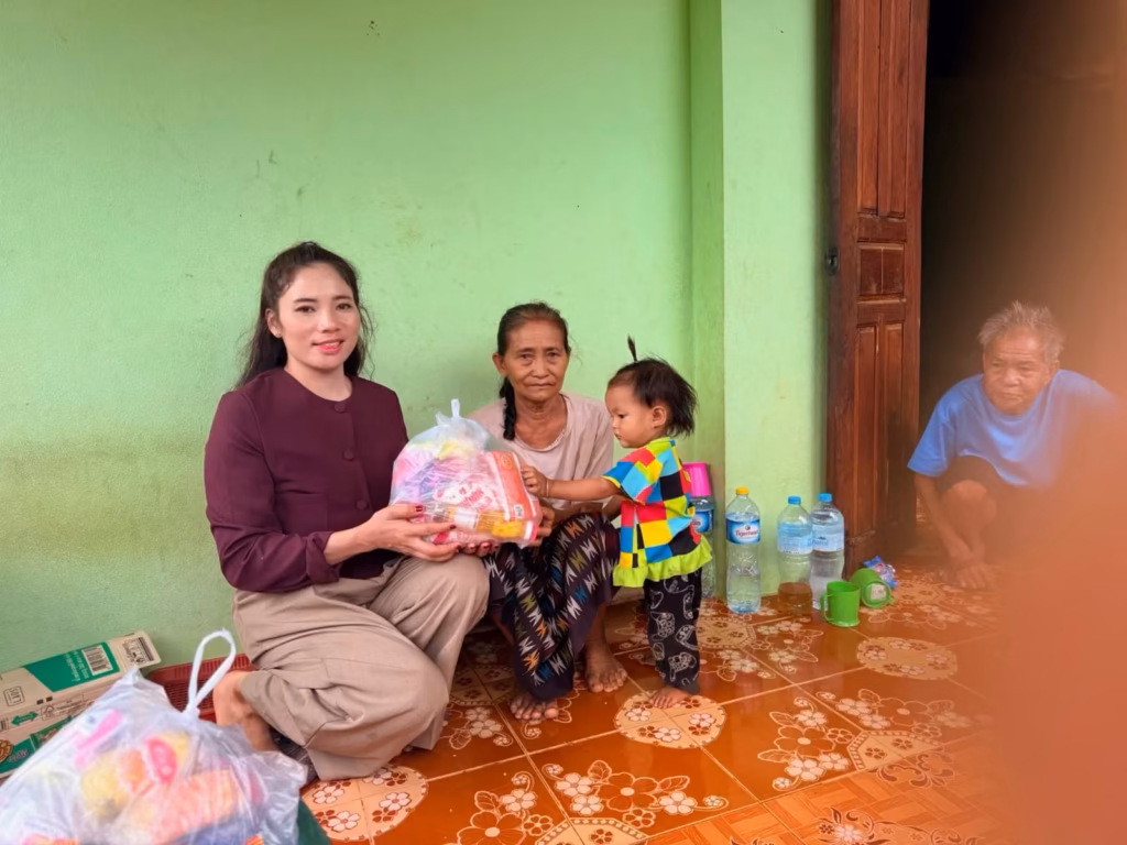 A ministry worker in a maroon top kneels to hand a food pack to a grandmother while her toddler reaches for it, an elderly man watching from the doorway.