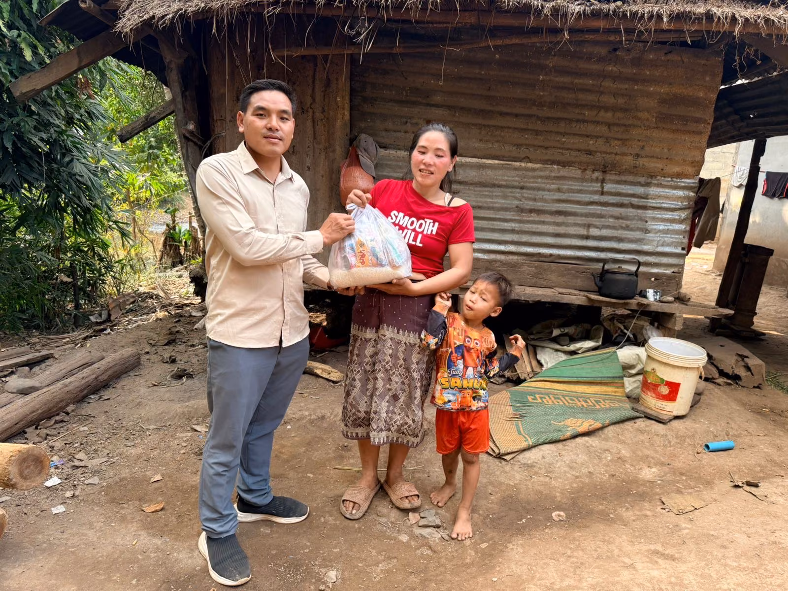 Pastor Khen passes a bag of rice and food to a young mother while her small son stands beside her outside their thatch-and-tin roofed home.