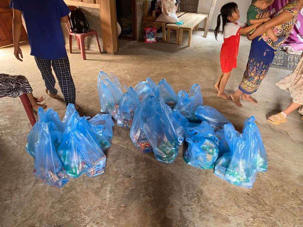 A pile of blue plastic bags filled with goods on a concrete floor while a child and a woman holding a toddler stand nearby in Laos.