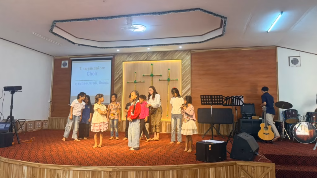 Children gathered on a carpeted church stage in front of three bamboo crosses with a woman among them; the screen reads Choir in Laos.