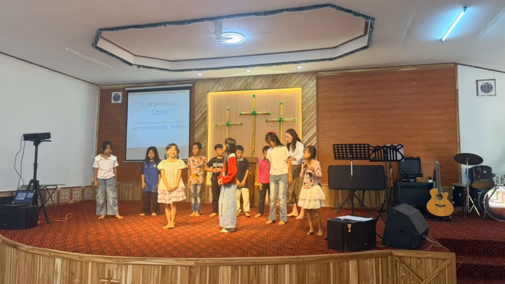 Children in a choir standing on a carpeted church stage with a woman beside them; the screen reads Choir in English and Lao in Laos.