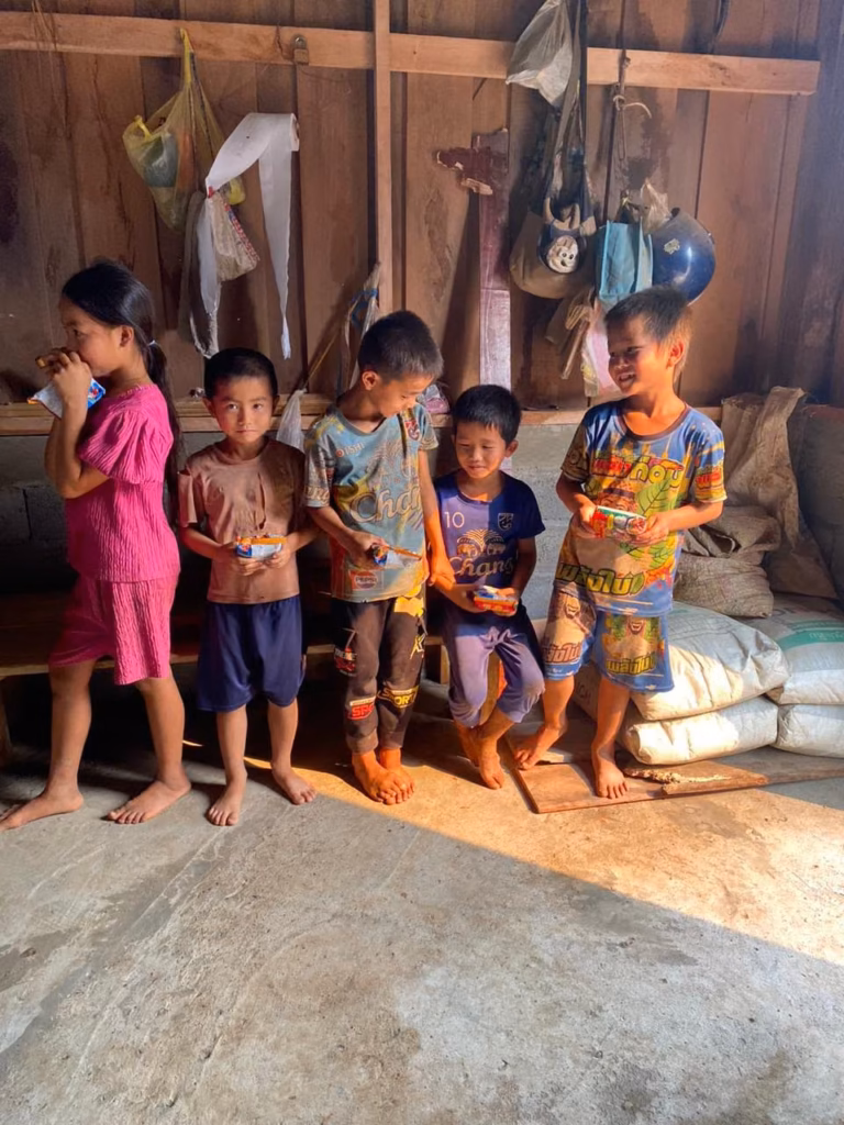 Five children standing against a wooden wall holding small packaged snacks, one of them drinking, inside a house in Laos.