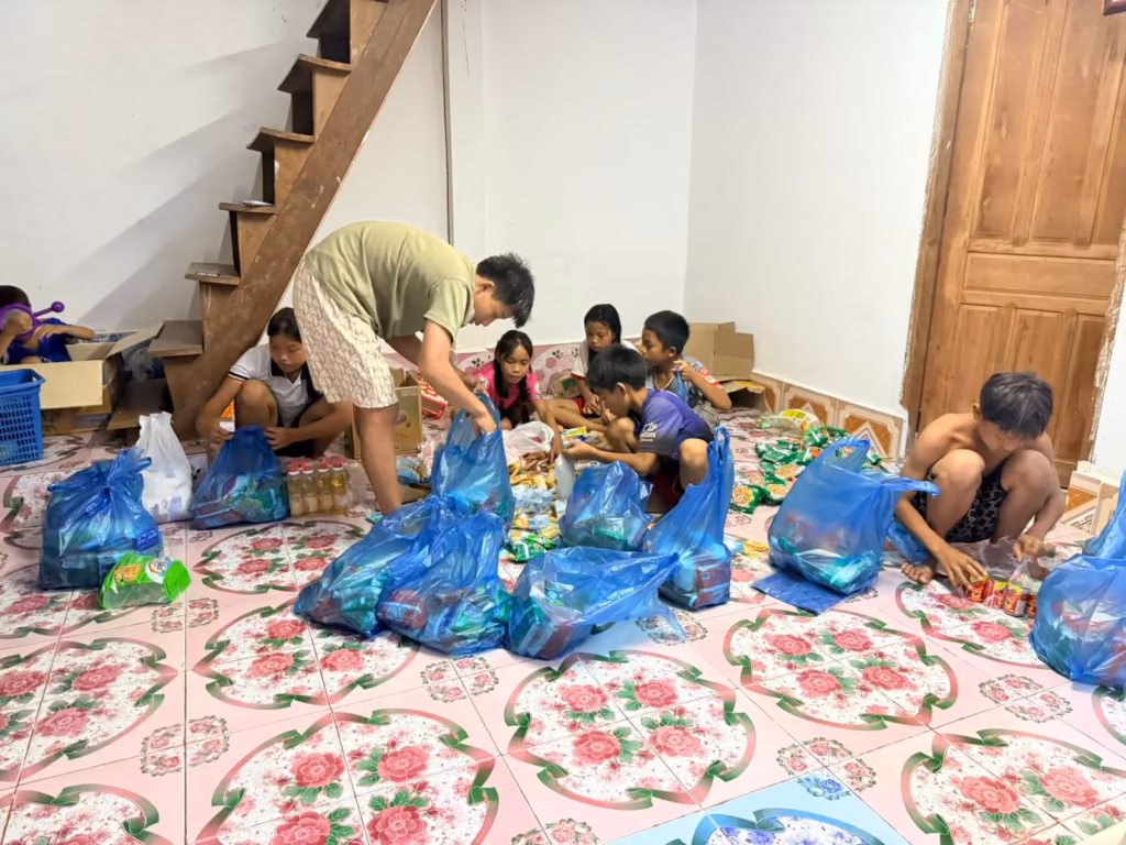 Children and a young man packing food and bottles of cooking oil into blue bags on a tiled floor in Laos.