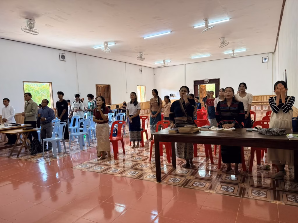A congregation standing among red and blue plastic chairs in a large hall with a tiled floor, many with hands raised to their faces, in Laos.