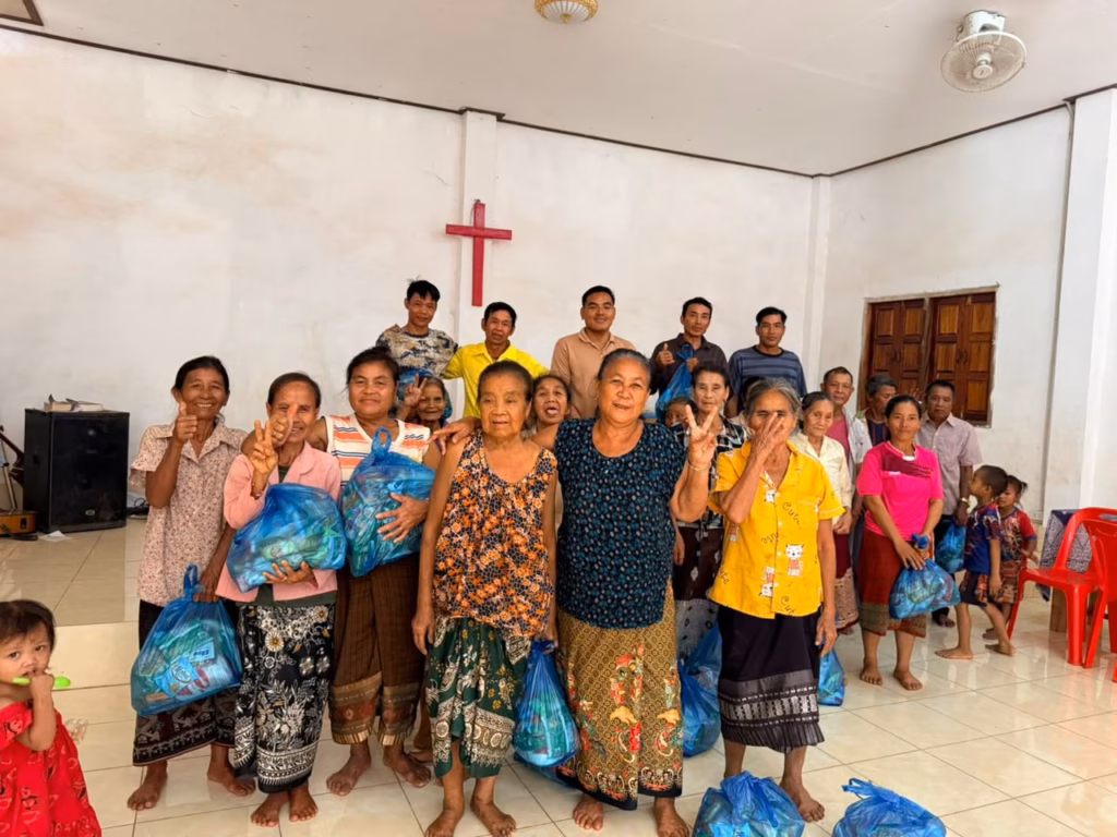 A group of women, men and small children posing in a church hall, many women holding blue bags of supplies and making peace signs in Laos. Pastor Khen is pictured.