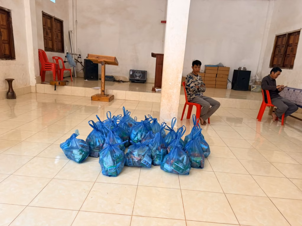 A pile of blue bags filled with food on the tiled floor of a church hall, with a wooden pulpit behind and two men seated on red chairs in Laos.