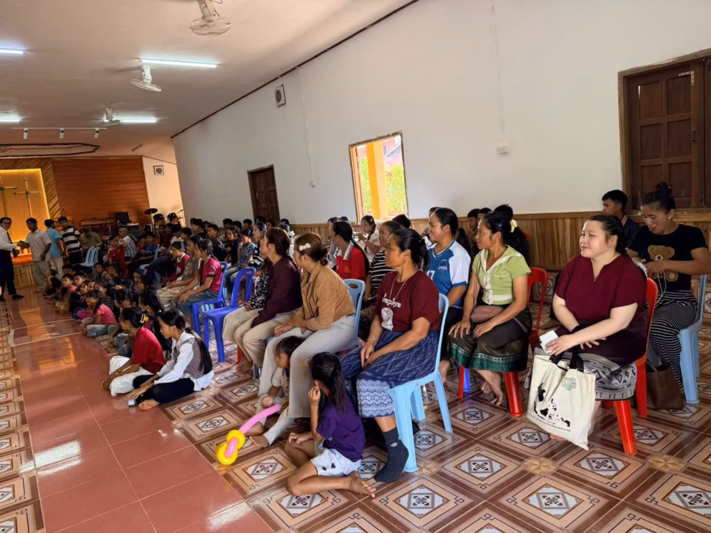 A pastor greeting a man at the left edge of a hall as women and children sit facing the stage in Laos.