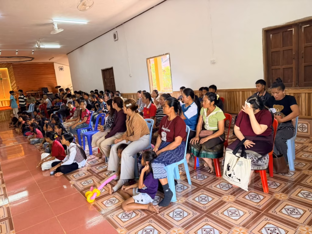 Women and children sitting on chairs and on a red tiled floor facing the stage of a large hall in Laos.