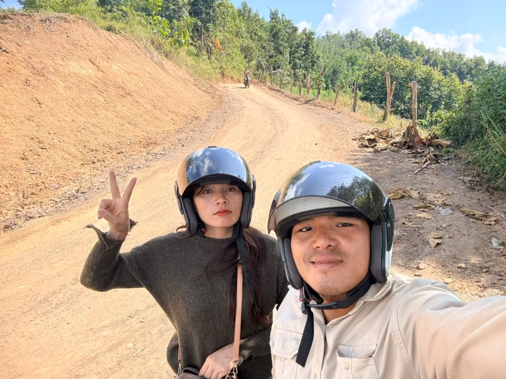 Pastor Khen and his wife, Mala, in black helmets taking a selfie on a dirt road beside a cut hillside, with Mala making a peace sign in Laos.