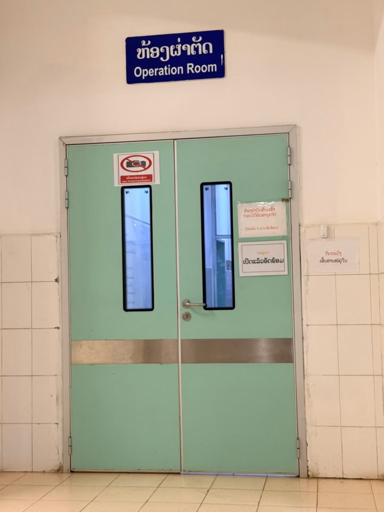 Closed green double doors under a blue sign reading Operation Room in English and Lao, with a no photograph notice, in the hospital hallway where a boy from Pastor Khen's orphanage had emergency surgery in Laos.