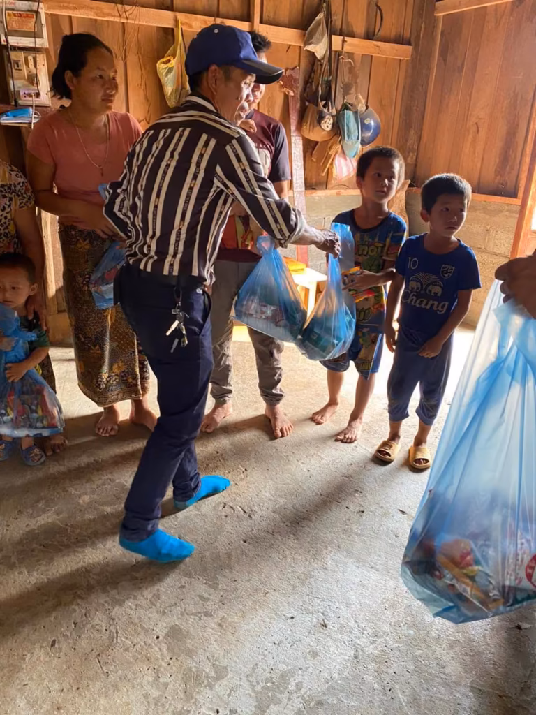 A pastor in a striped shirt and blue shoe covers handing blue plastic bags of goods to children inside a wooden house in Laos.