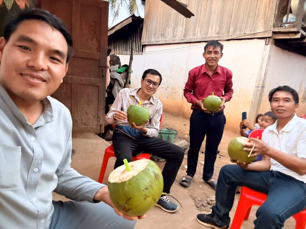 Four men holding green coconuts with straws in a dirt yard beside village houses, with two small children behind them in Laos.
