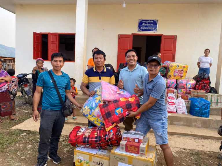 Pastor Khen, a fellow pastor and two other men holding blankets and a pillow in front of stacked bedding and boxes outside a village office in Laos.