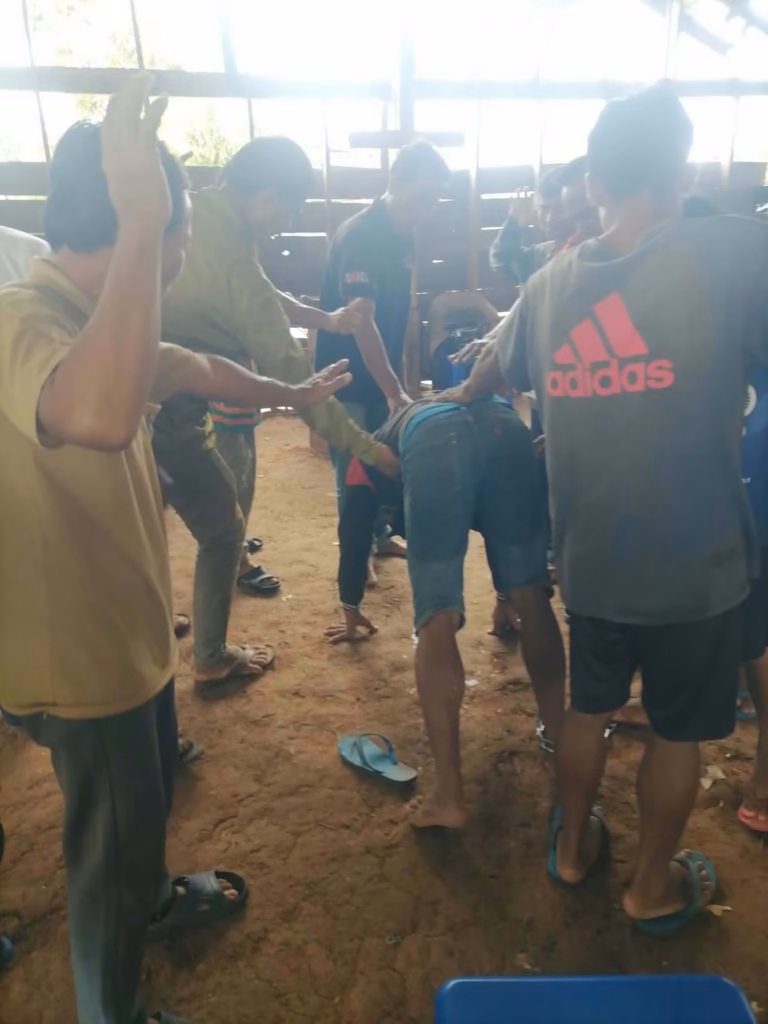 A pastor and several other men lay their hands on a man bending forward on a dirt floor inside a wooden building in Laos.