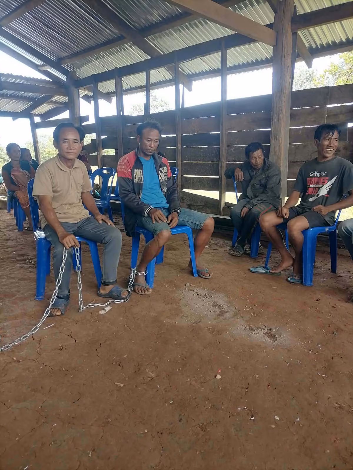 Four men sit on blue plastic chairs under a metal-roofed wooden shelter, with chains running from the ankles of two of them across the dirt floor in Laos.