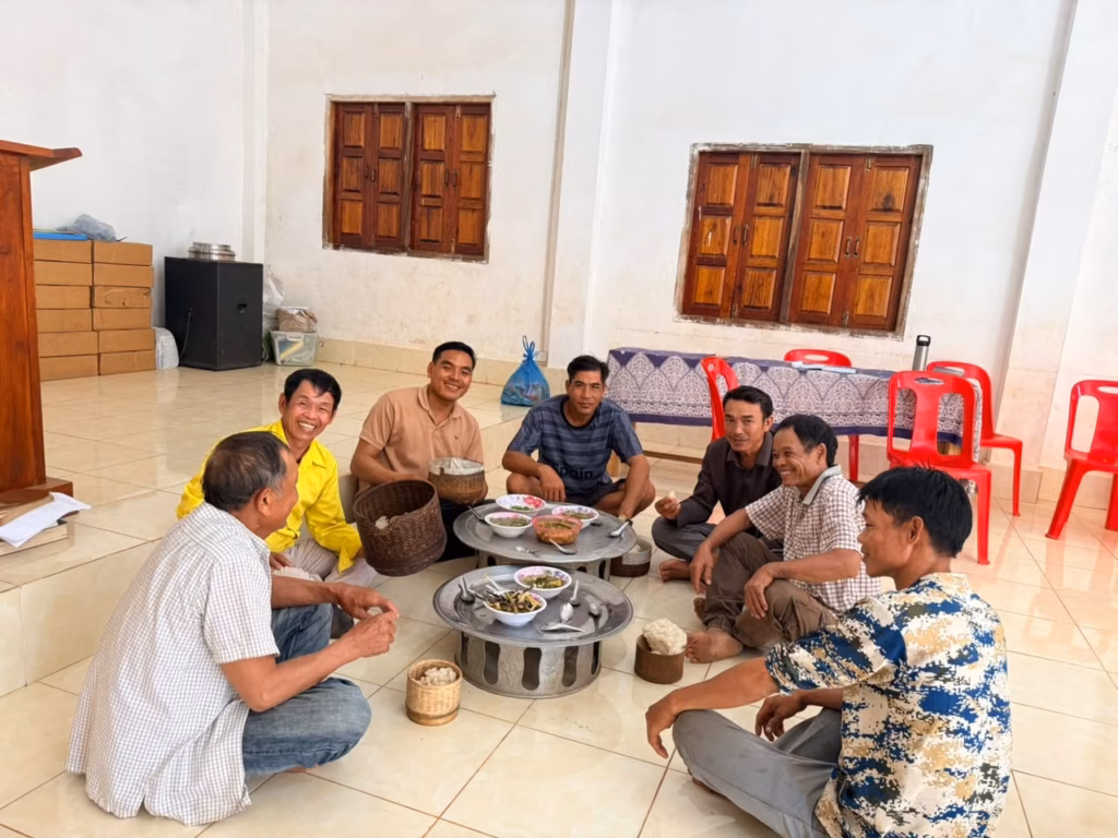 Seven men sitting on a tiled floor in a church hall sharing a meal of sticky rice and dishes from two metal trays in Laos. Pastor Khen is pictured.
