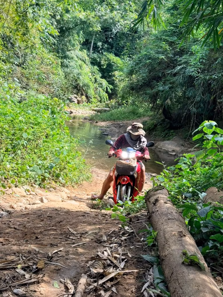 A rider in a bucket hat and face cover on a red motorbike climbing a dirt path beside a stream in a green forest in Laos.