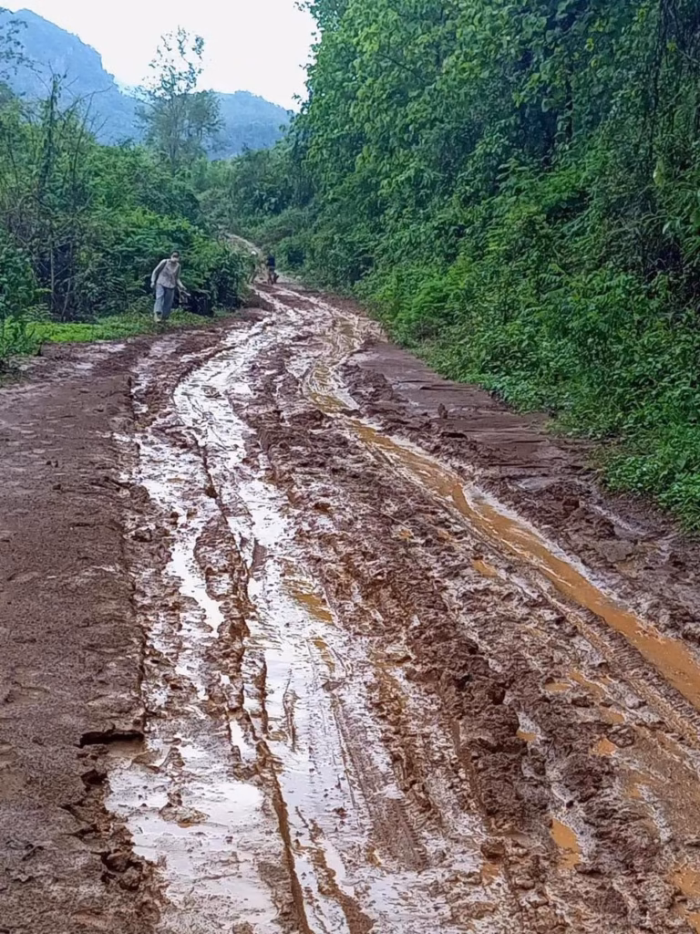 A muddy, rutted dirt road winding between green forested hills, with a person walking and a motorbike far ahead in Laos.