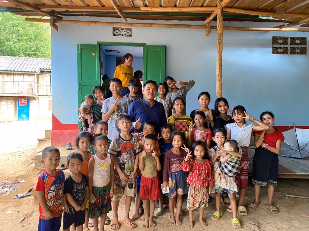 Pastor Khen in a dark blue shirt giving a thumbs up in the middle of many smiling children outside a blue house with green shutters in Laos.