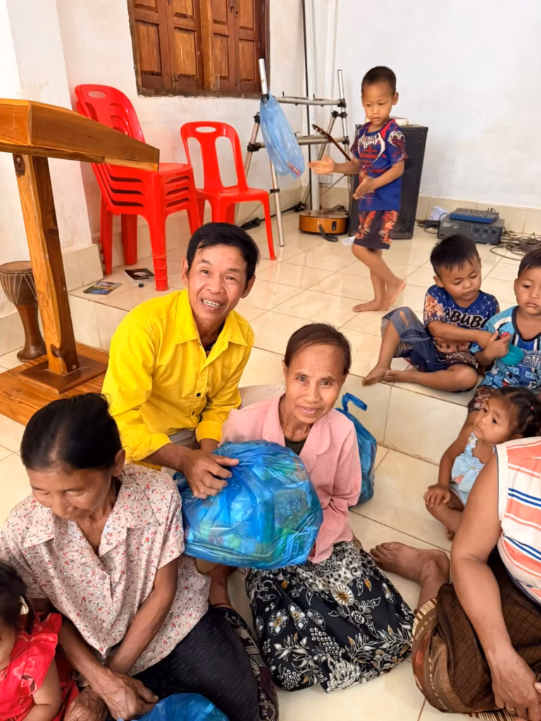 A pastor in a yellow shirt kneeling to hand a blue bag of food to a seated older woman, with children and other women on a tiled church floor in Laos.