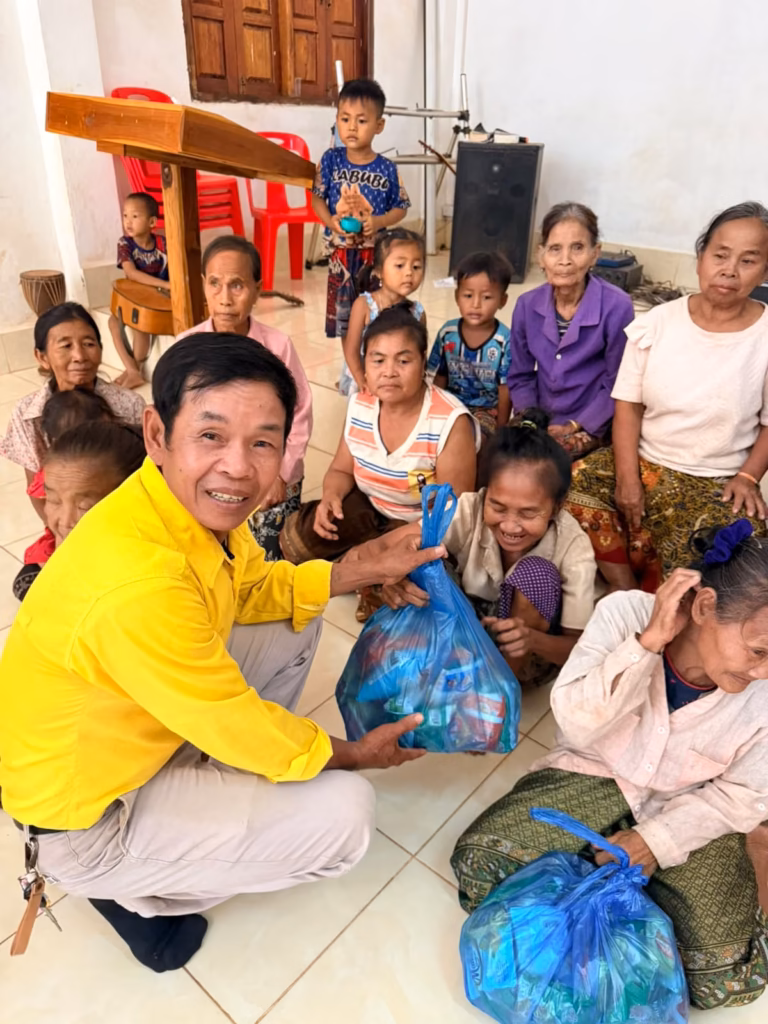 A pastor in a yellow shirt crouching to hand a blue bag of food to a smiling woman, with a group of women and children seated around them in Laos.