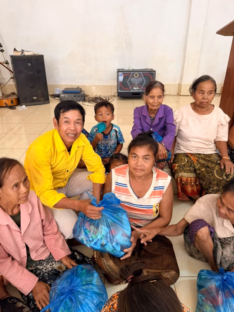 A pastor in a yellow shirt handing a blue bag of food to a woman in a striped top, surrounded by seated women and a small child in a church hall in Laos.