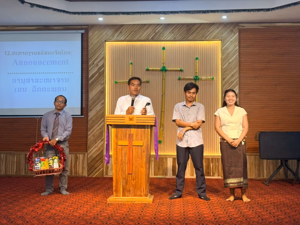 Pastor Khen speaking at a wooden pulpit during the announcement time while a man holds a gift basket and a man and woman stand beside him; the slide reads Announcement in English and Lao.