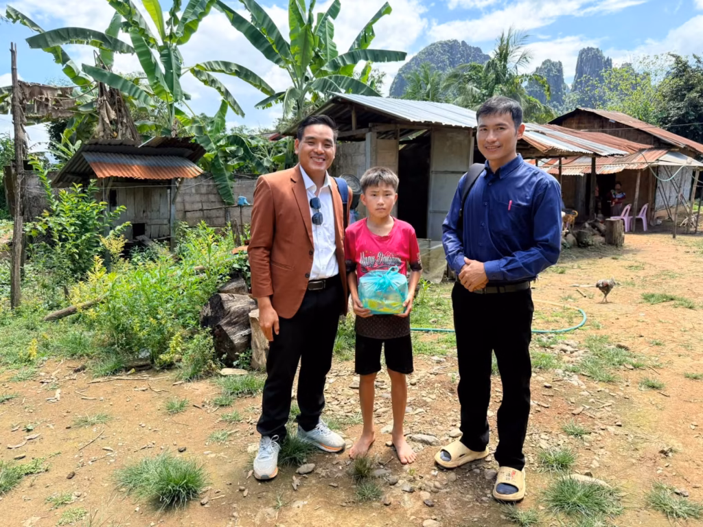 Pastor Khen and a fellow pastor standing with a barefoot boy holding a gift bag in a Laos village below limestone karst mountains.