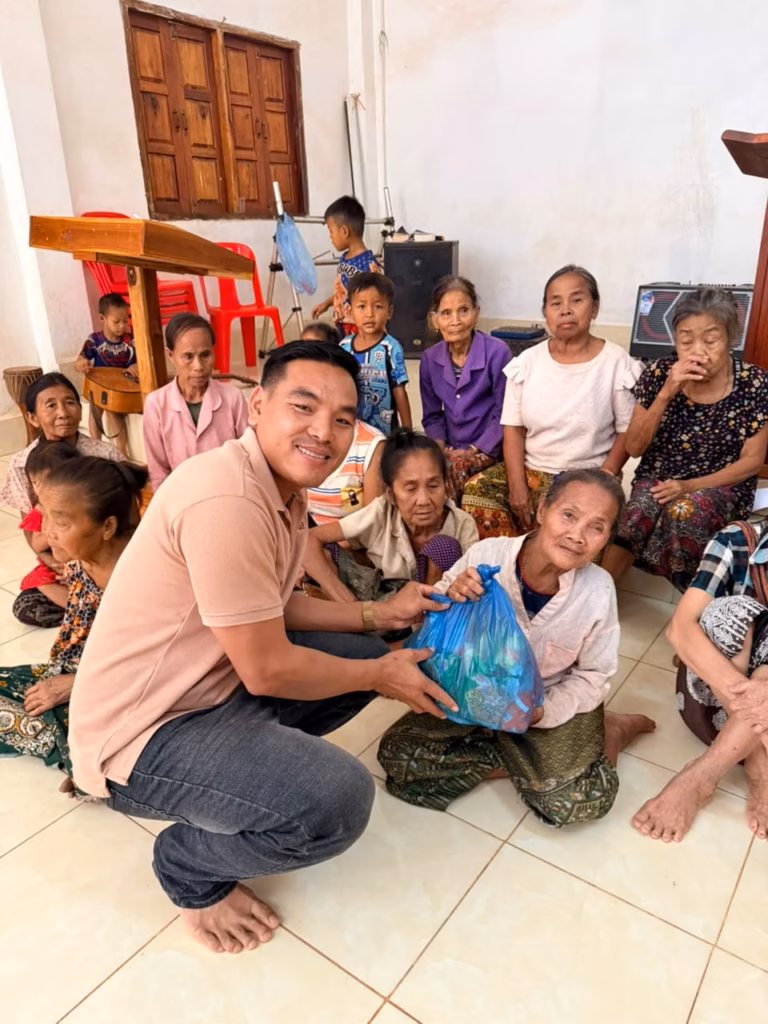 Pastor Khen crouching to hand a blue bag of supplies to an elderly woman seated among other women and children in a church hall in Laos.