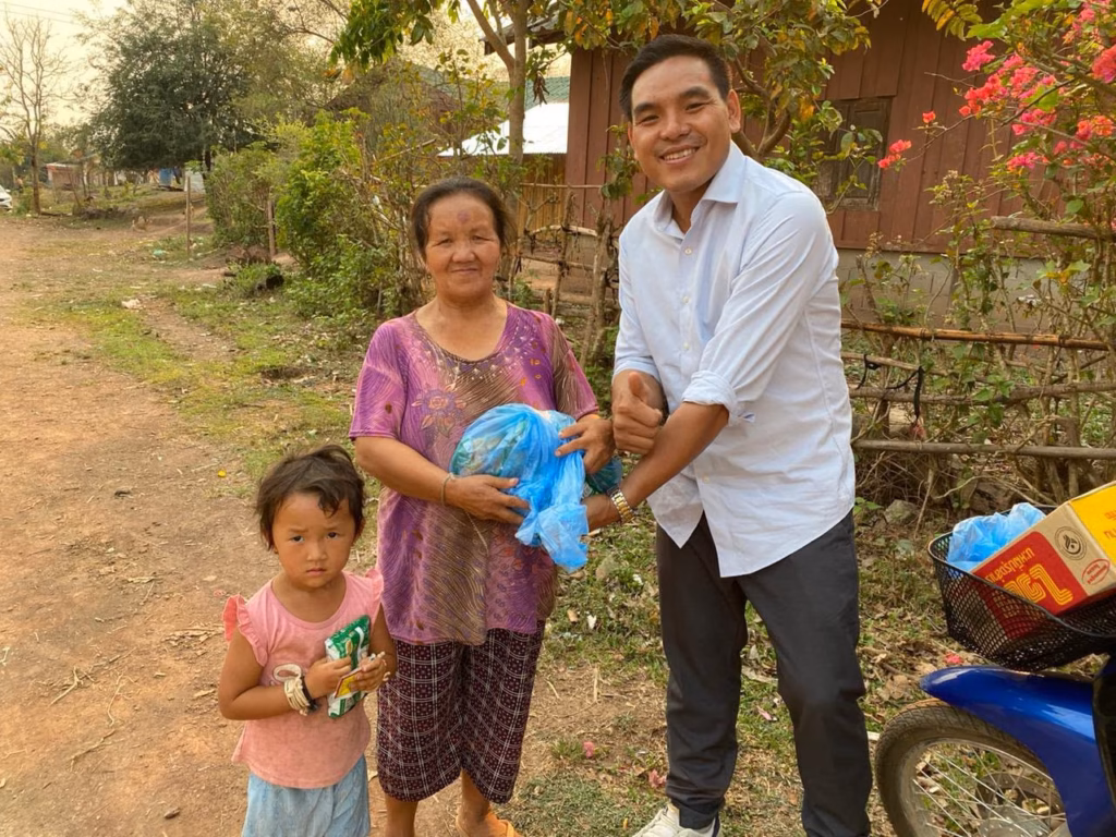 Pastor Khen giving a blue bag of goods to an older woman, with a young girl holding a snack beside them on a village road in Laos.