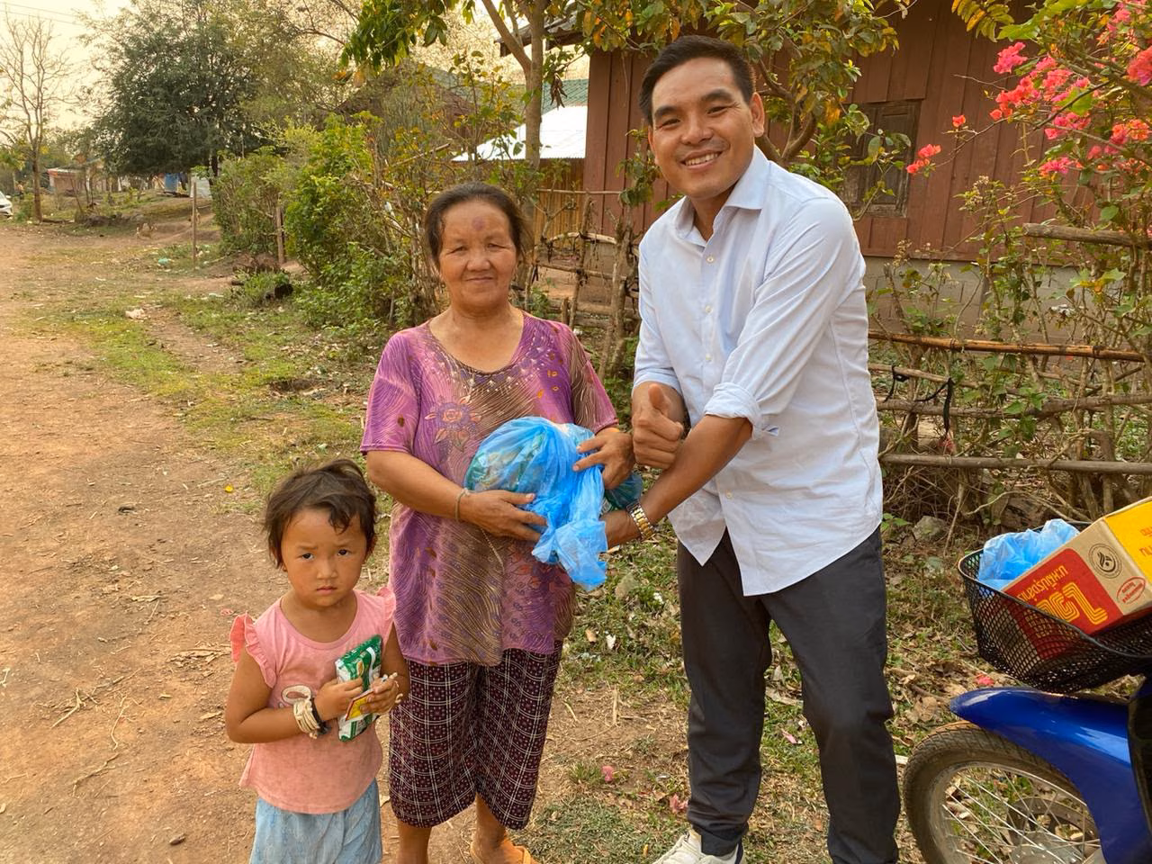 Pastor Khen giving a blue bag of goods to an older woman, with a young girl holding a snack beside them on a village road in Laos.