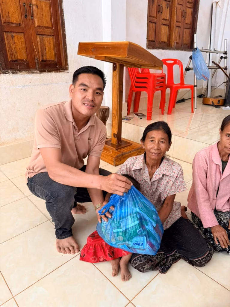 Pastor Khen crouching beside a wooden pulpit and handing a blue bag of supplies to a woman holding a small child in a red dress in Laos.