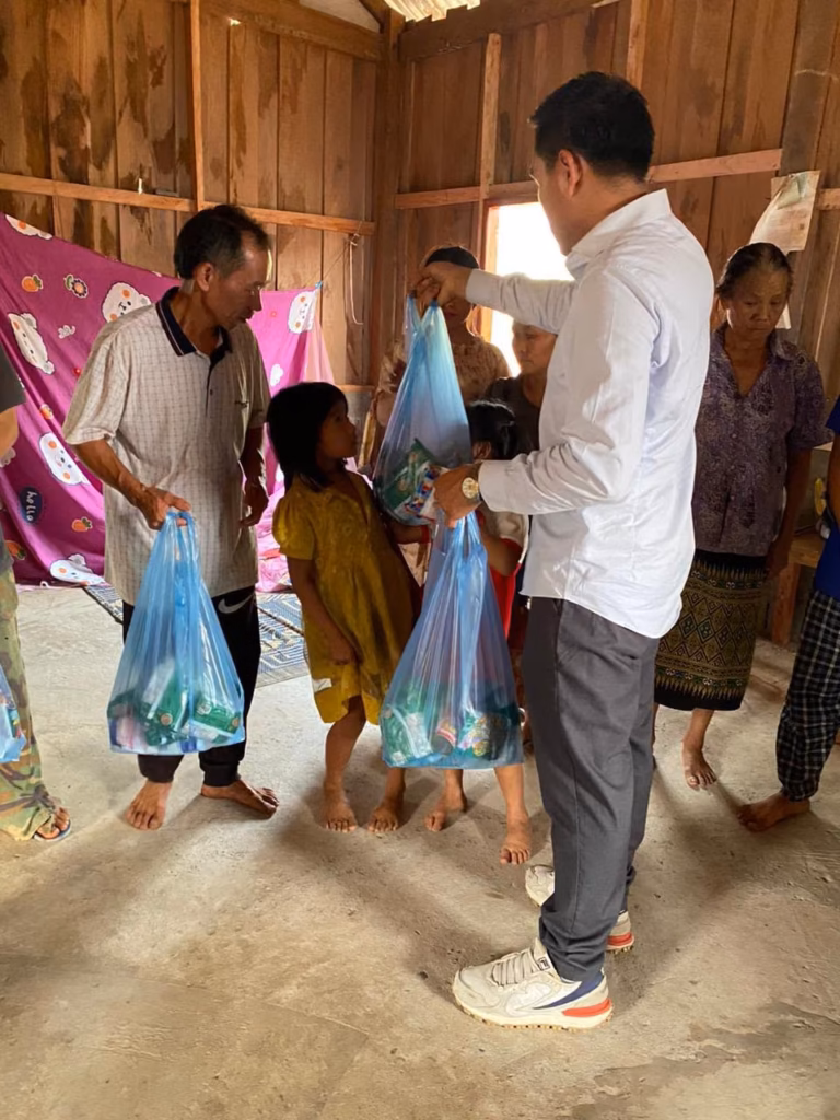 Pastor Khen in a white shirt handing blue gift bags to a girl and an older man inside a wooden house in Laos.