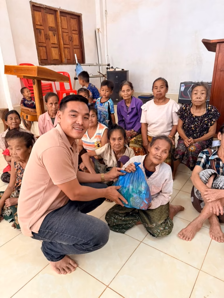 Pastor Khen crouching on a tiled floor and holding out a blue bag of supplies to a seated elderly woman, with a crowd behind her in Laos.