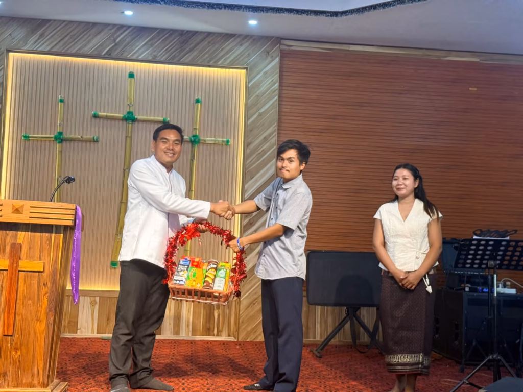 Pastor Khen shaking hands with a young man while they hold a red tinsel gift basket of snacks on a church stage in Laos, with a woman standing nearby.