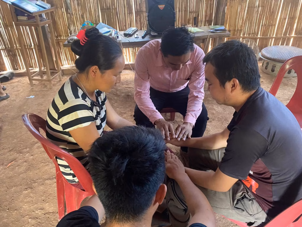 Pastor Khen seated and praying with hands joined with a woman and a man in a bamboo house, with another man's head in the foreground in Laos.