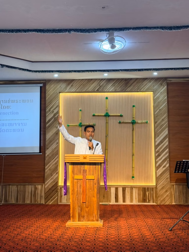 Pastor Khen speaking into a microphone with one arm raised at a wooden pulpit in front of three bamboo crosses, with a Lao slide beside him in Laos.