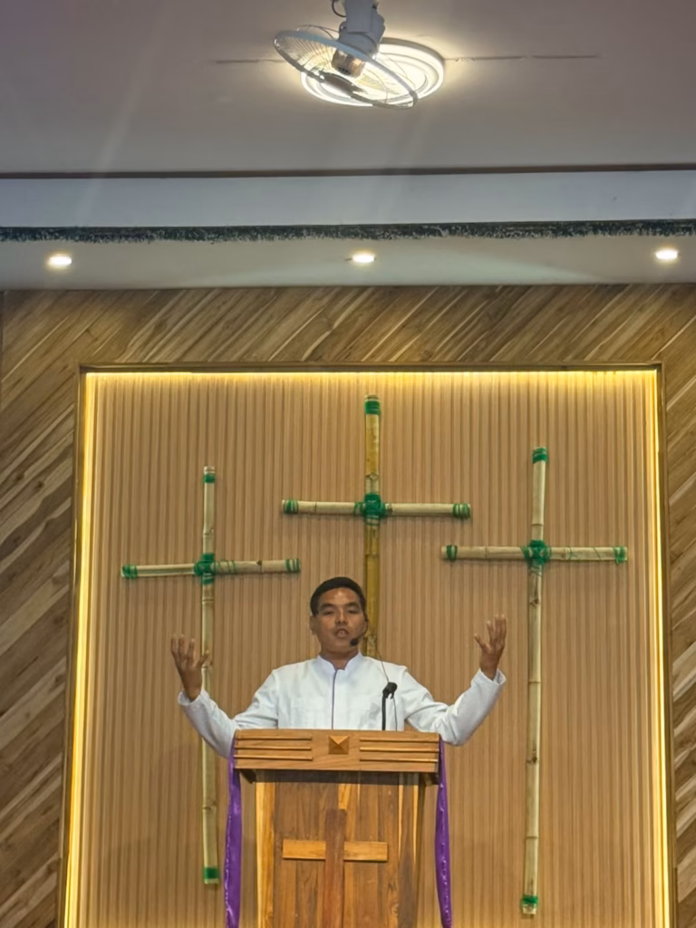 Pastor Khen preaching with both arms raised behind a wooden pulpit in front of three bamboo crosses in Laos.