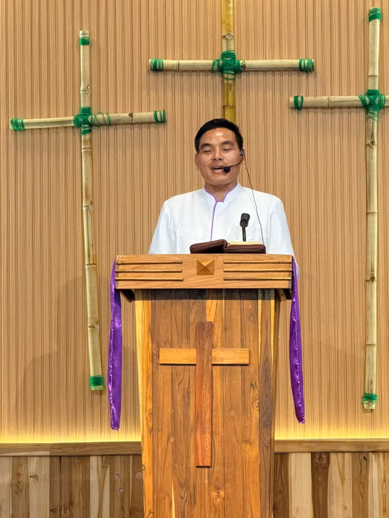 Pastor Khen speaking with eyes closed at a wooden pulpit with an open Bible, bamboo crosses on the wall behind him in Laos.