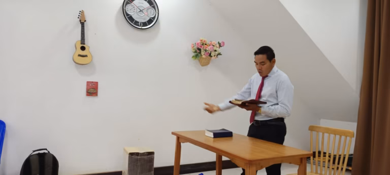 Pastor Khen preaching with an open Bible in his hand beside a wooden table in a white room with a ukulele on the wall in Laos.