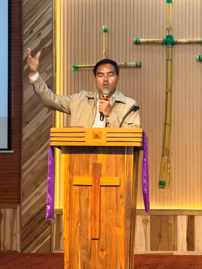 Pastor Khen preaching with one hand raised and a microphone in the other at a wooden pulpit in front of bamboo crosses in Laos.