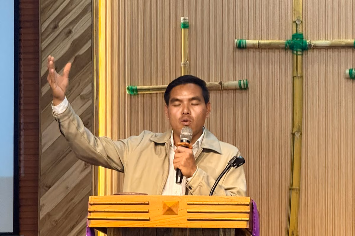Pastor Khen preaching with one hand raised and a microphone in the other at a wooden pulpit in front of bamboo crosses in Laos.