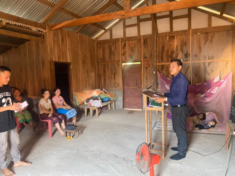 Pastor Khen preaching from a small wooden lectern inside a wooden house while women sit on plastic chairs and a man holds papers in Laos.