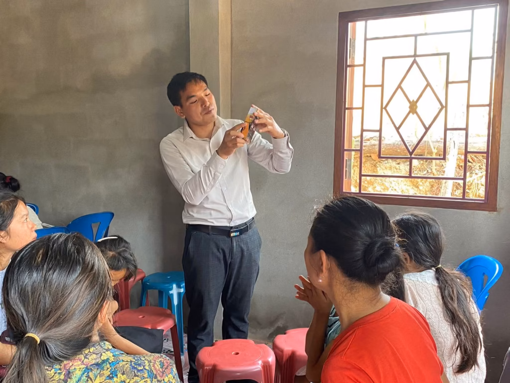 Pastor Khen holding a small booklet in both hands while teaching women seated on plastic stools in a concrete room in Laos.