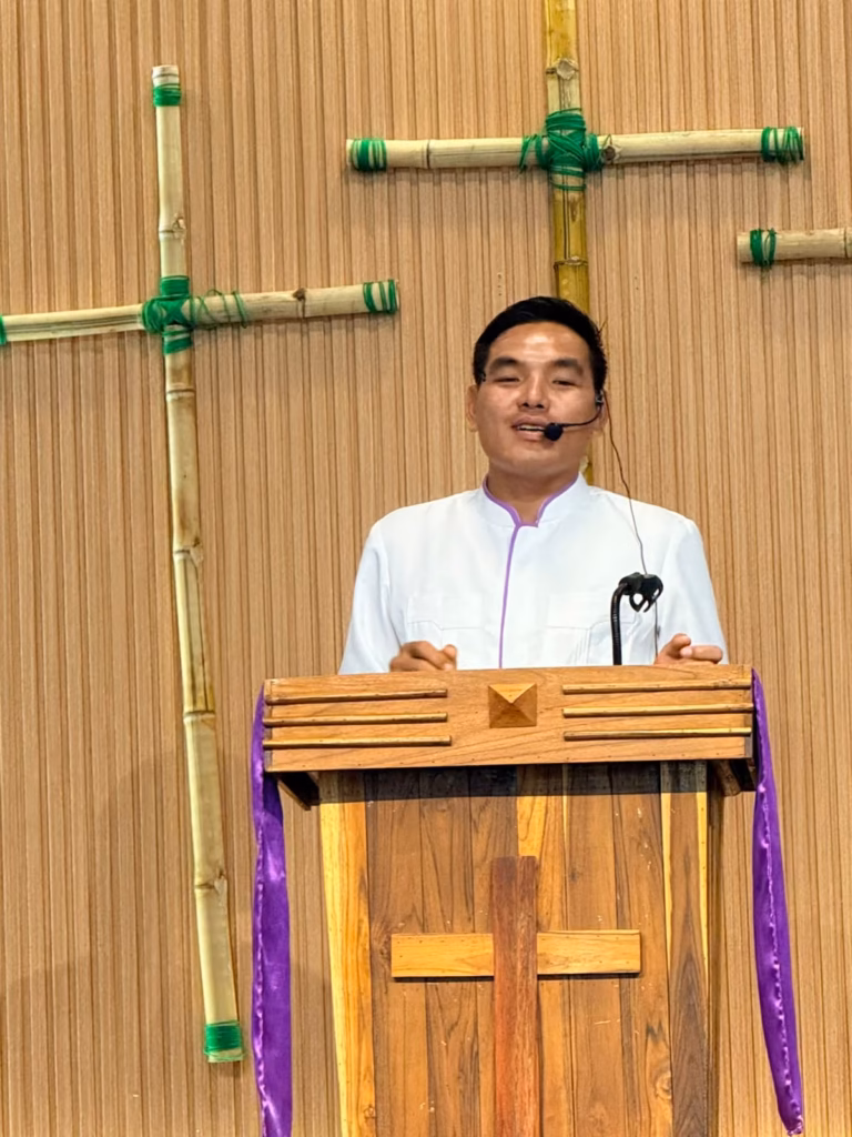 Pastor Khen speaking into a headset microphone at a wooden pulpit with a purple cloth and bamboo crosses behind him in Laos.