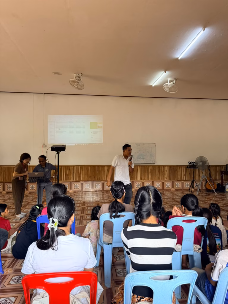 Pastor Khen speaking into a microphone in front of seated children while two people work at a laptop and projector in Laos.