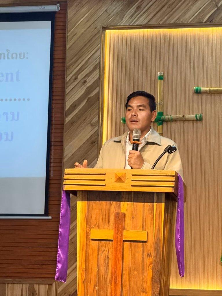 Pastor Khen speaking into a microphone at a wooden pulpit with a cross, beside a projector screen showing Lao text in Laos.