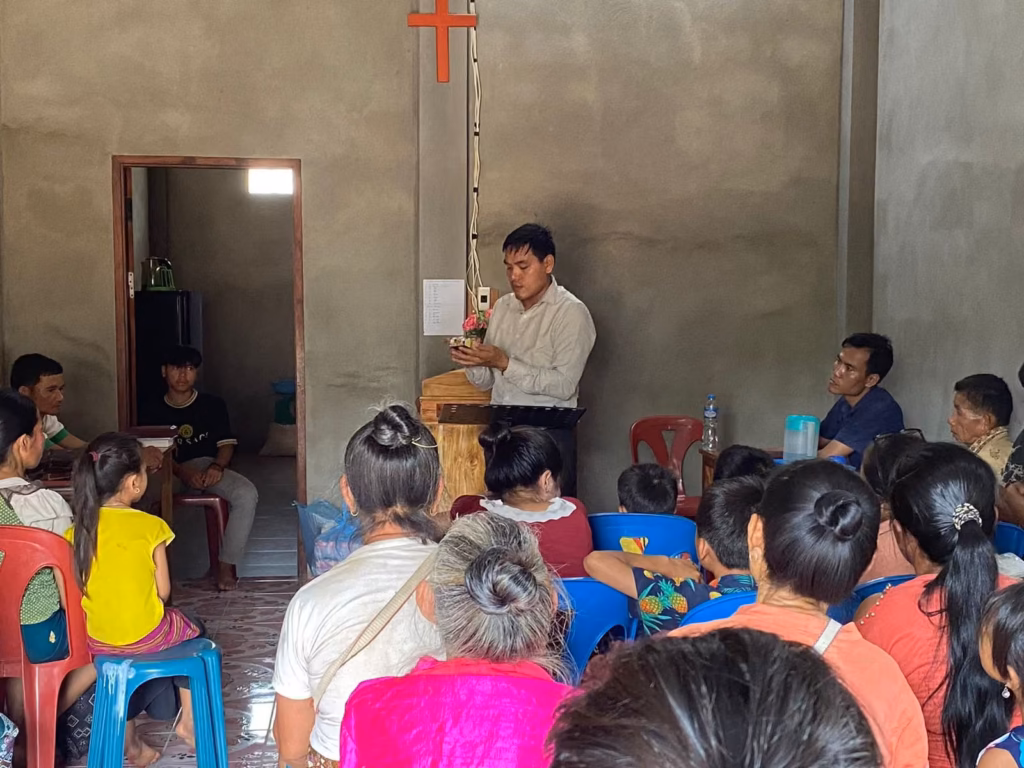 Pastor Khen speaking at a wooden lectern in a concrete room with a red cross, while a seated congregation faces him in Laos.