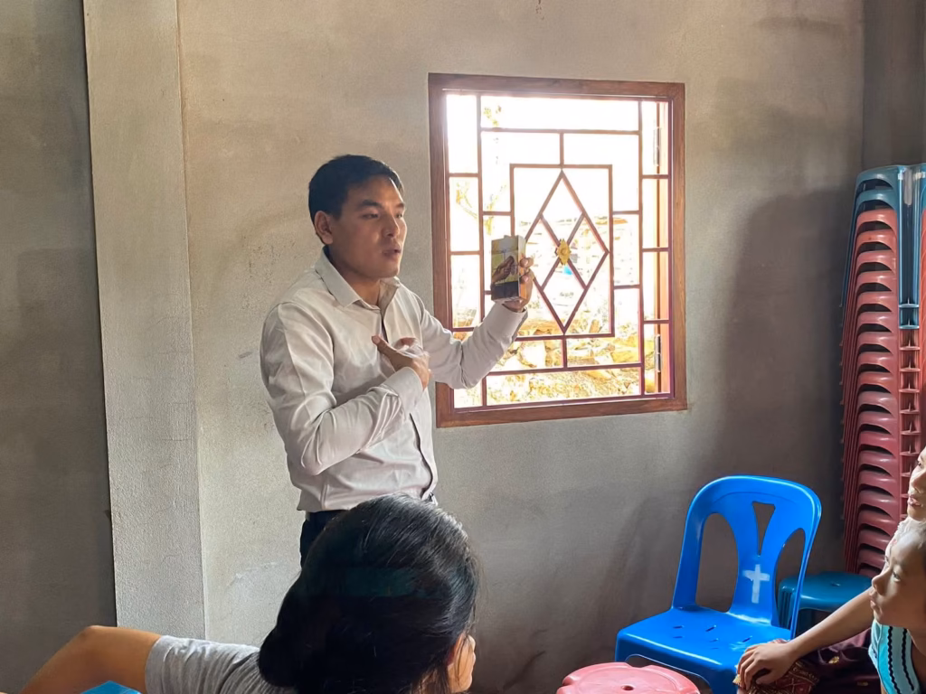 Pastor Khen holding up a small booklet and touching his chest while teaching beside a window in a concrete building in Laos.