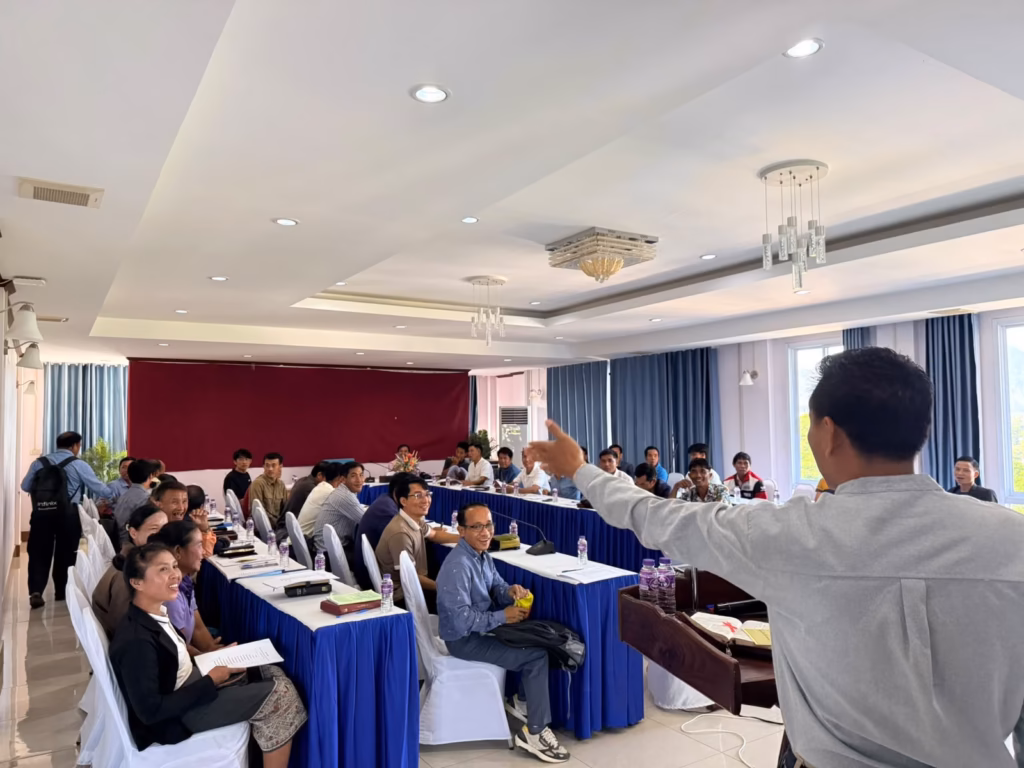 Pastor Khen teaching with an outstretched arm beside a wooden pulpit to smiling men and women seated at blue-draped tables at the pastors' conference in Vang Vieng, Laos.